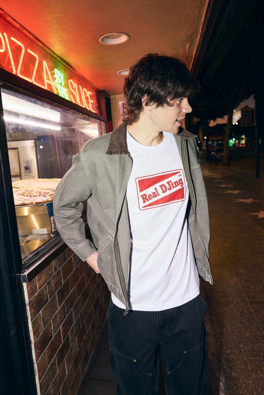 Model in white Real DJing tee under a grey jacket, lit by red pizza-shop neon