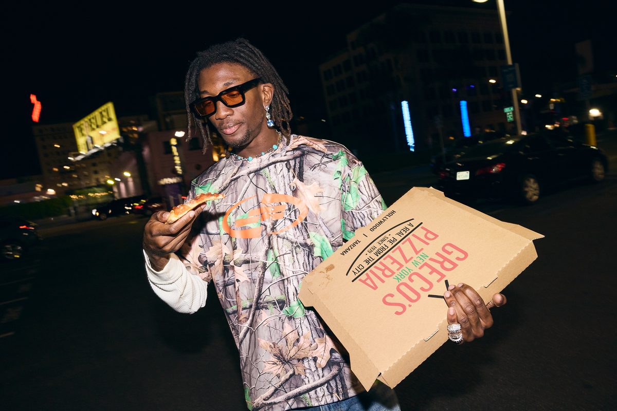 Solo model eating pizza on a Hollywood street at night, wearing the Realtree camo long-sleeve with the orange wave chest graphic